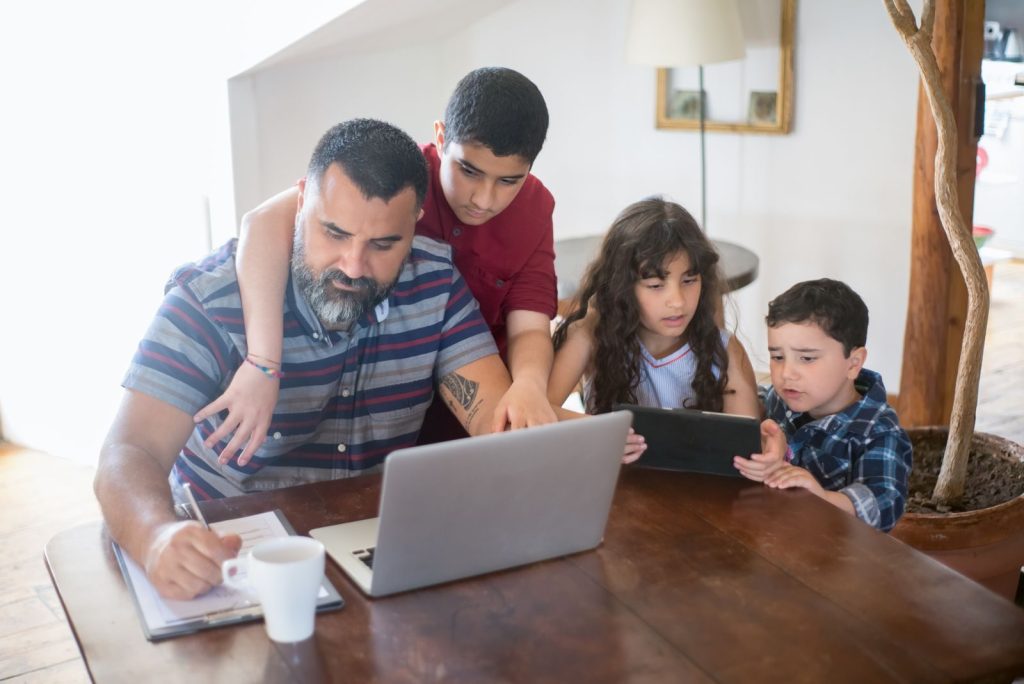 A father and his children engaged in activities with a laptop and tablet at home.