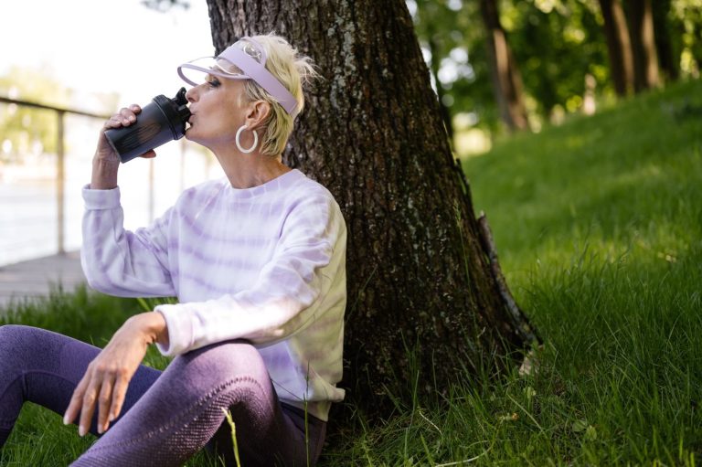 Elderly woman sitting by a tree in a park, sipping water under the sun.