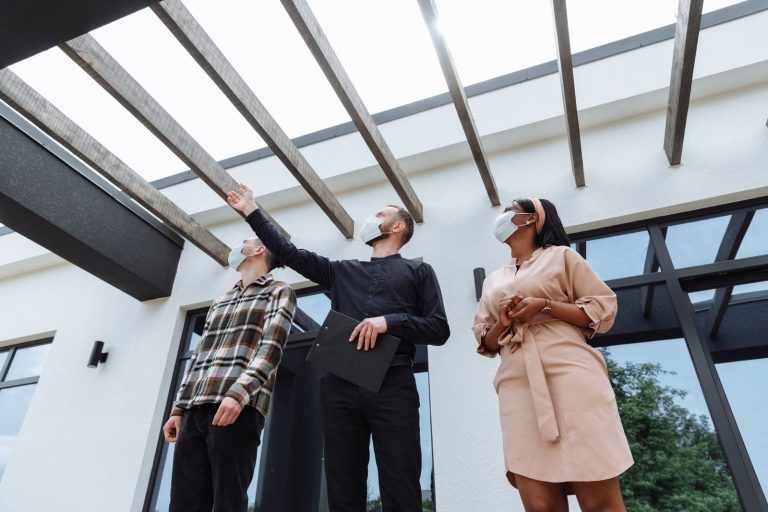 Masked real estate agent showcases modern architecture to clients outdoors.