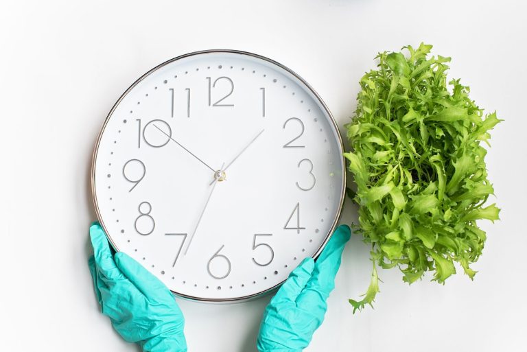White wall clock held by gloved hands beside lush green plant on white background.