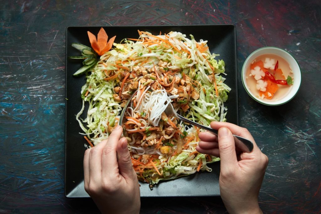 Top view of hands holding fork and spoon with noodles and fresh vegetables on plate.