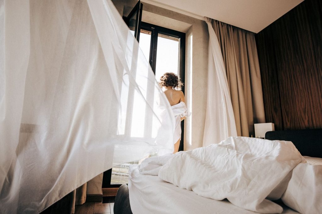 A person stands by an open window in a bedroom with flowing curtains and soft morning light.