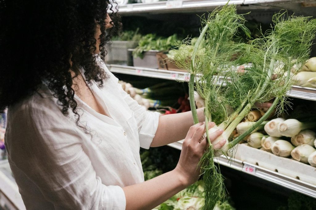 A woman in a white top choosing organic fennel in a grocery store produce section.