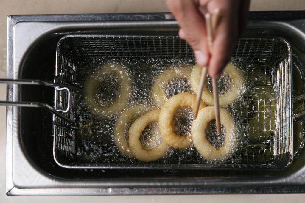 Overhead view of onion rings frying in hot oil with chopsticks in a deep fryer.