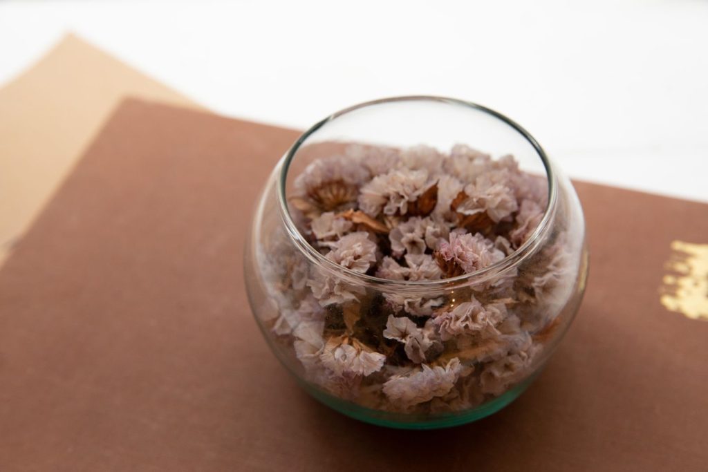 A close-up of delicate dried flowers arranged in a transparent glass bowl on a textured surface.