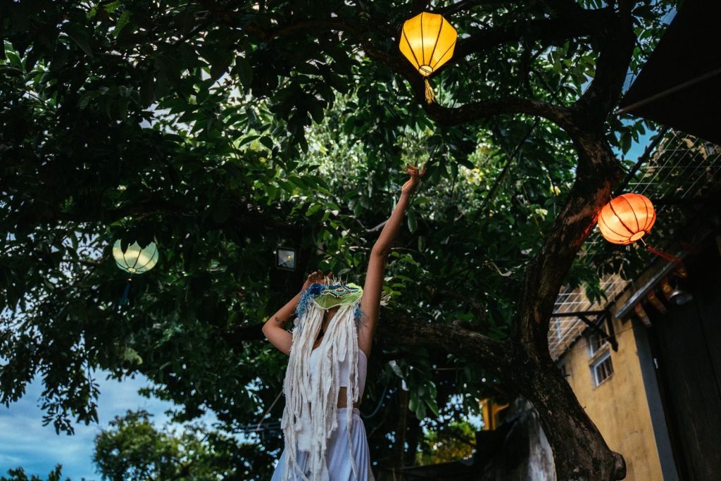 A woman with a mask raises her hand towards colorful lanterns in a lush tree at a Taiwanese festival.