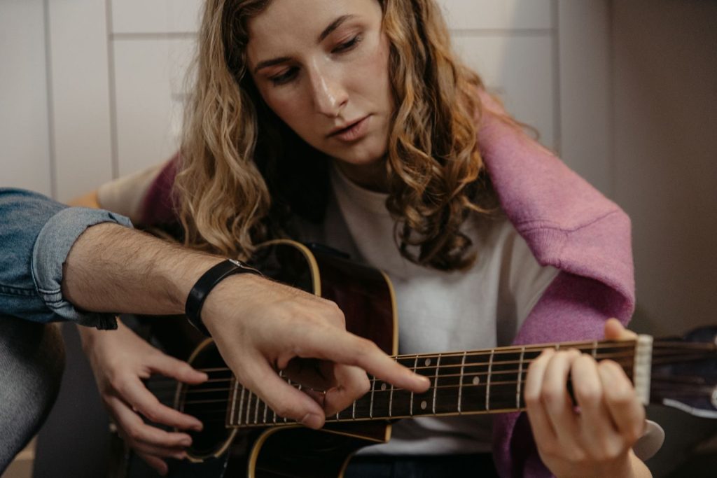 A woman is learning to play acoustic guitar with guidance indoors, focusing on finger placement.