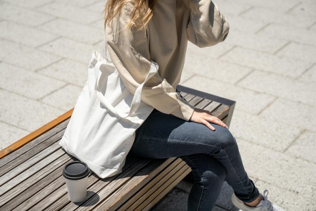 Person sitting on a bench with coffee wearing denim jeans and holding a tote bag.