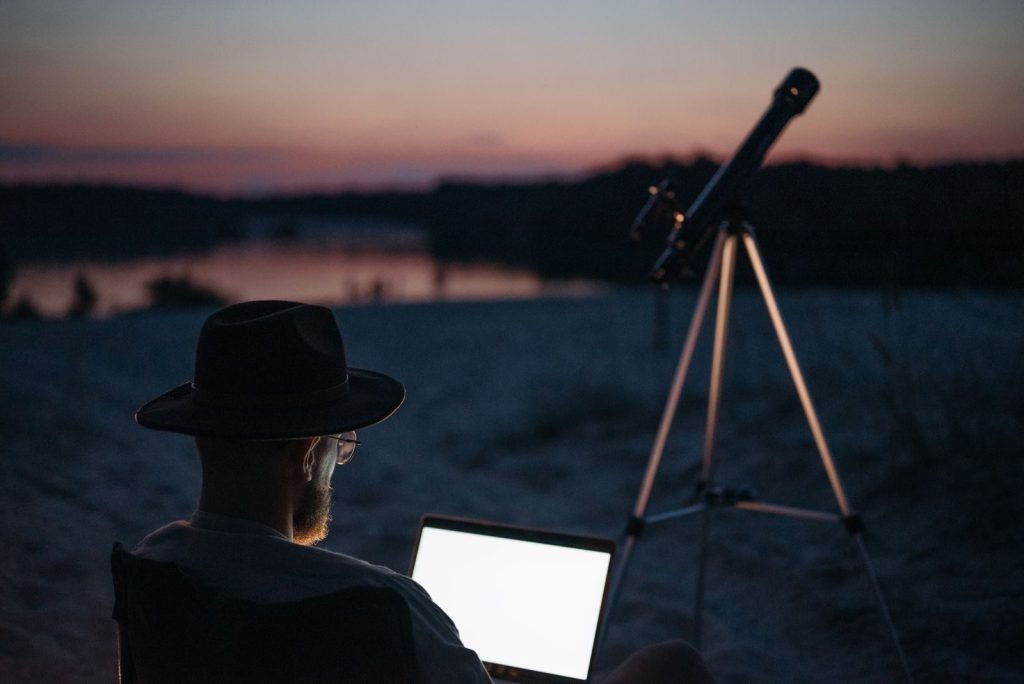 A man enjoying stargazing by a serene lake at twilight, using a telescope and laptop.