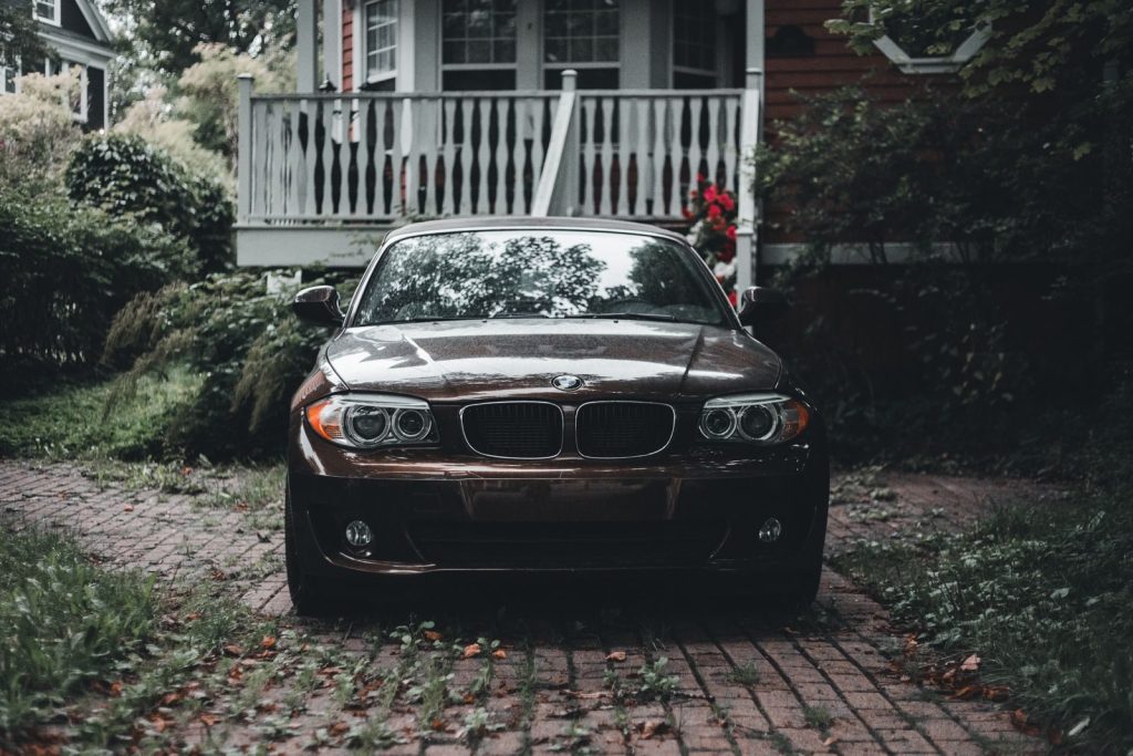 A black sedan parked on a brick paved path, surrounded by greenery and a wooden house.