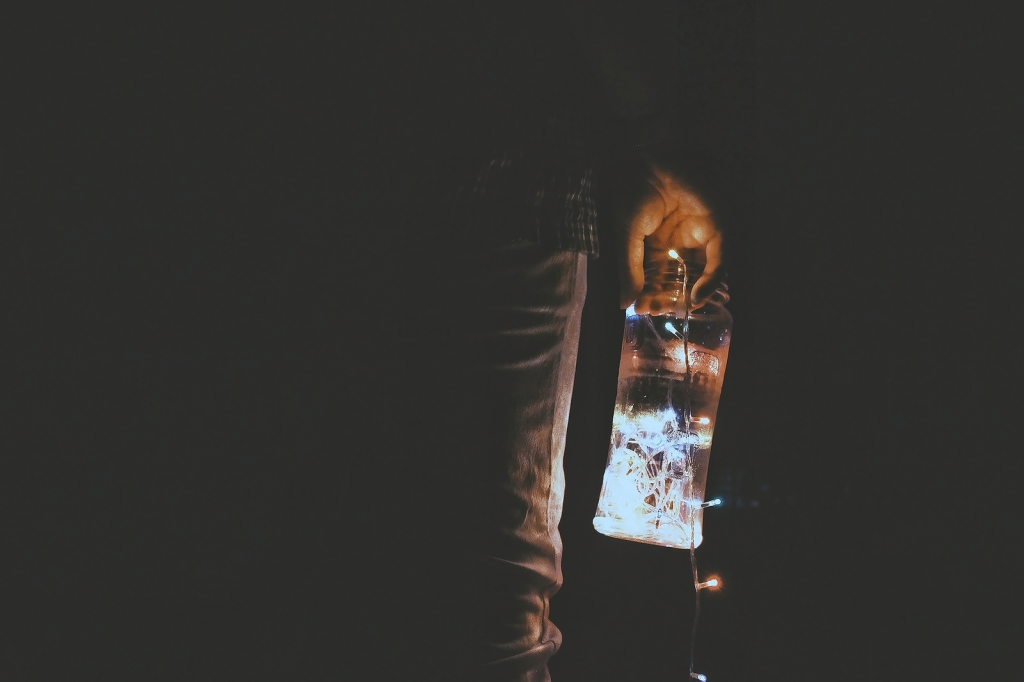 A hand holding a lighted bottle with string lights in a dark setting, creating a mysterious mood.