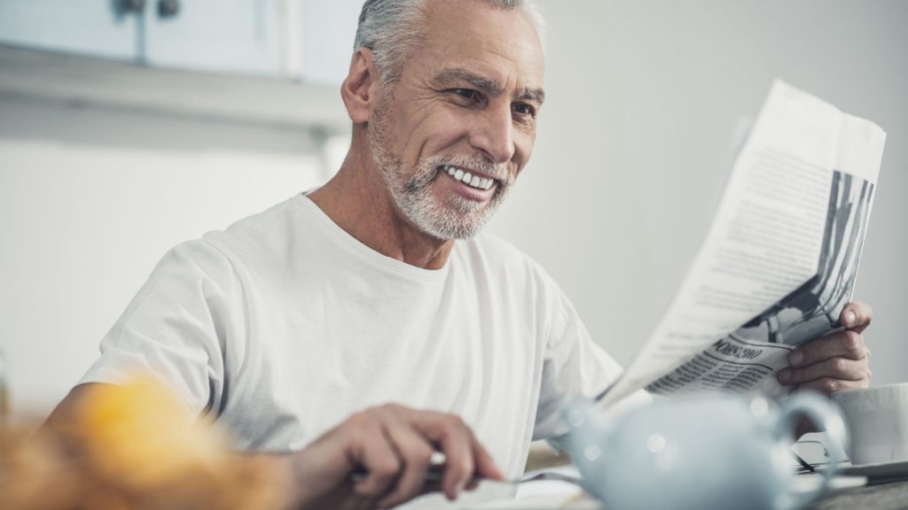 Morning at home. Beaming wrinkled elderly man enjoying his morning at home while eating crepes and reading newspaper