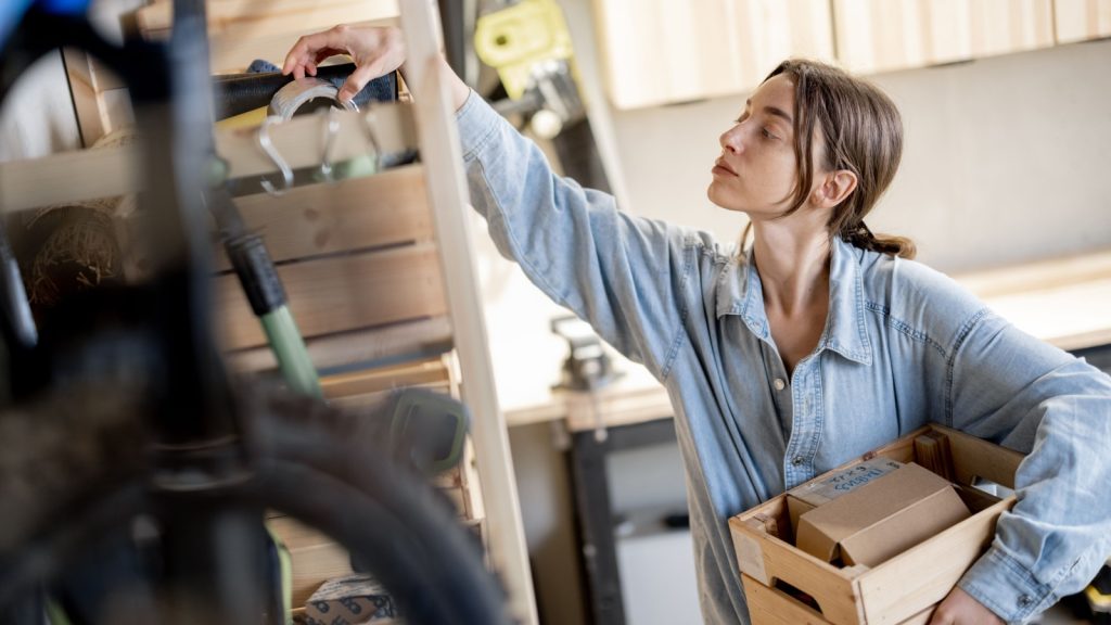 Young handywoman searching some working tools on a wooden shelves in the workshop. Concept of organization in home workshop or storage