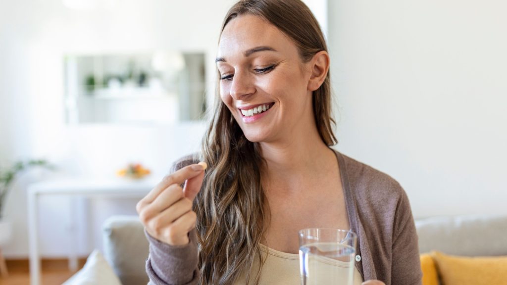 Head shot portrait happy woman holds pill glass of water, takes daily medicine vitamin D, omega 3 supplements, skin hair nail strengthen and beauty, medication for health care concept