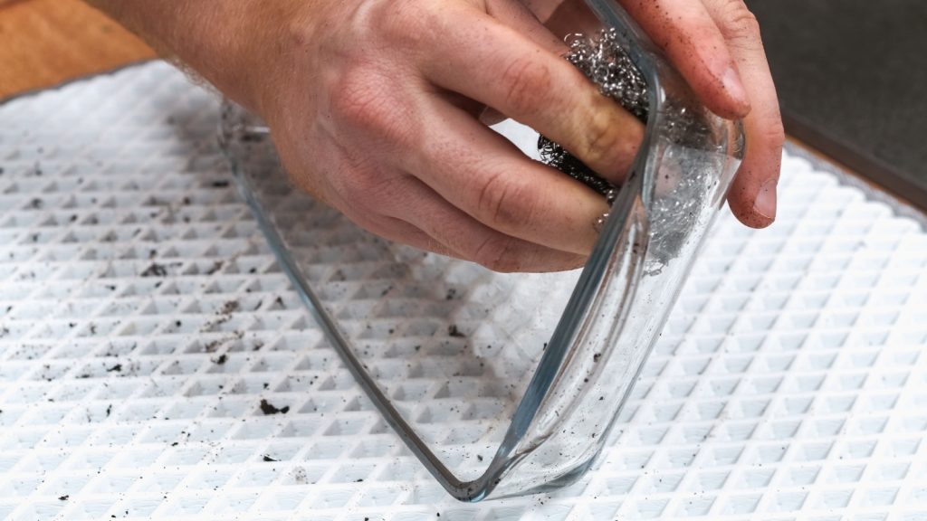 A man is cleaning a dirty baking dish with a thick layer of carbon with an iron washcloth. Glassware for baking with soot, carbon deposits, old dried fat.
