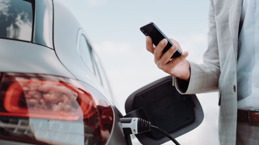 Man holding smartphone while charging car at electric vehicle charging station, closeup.