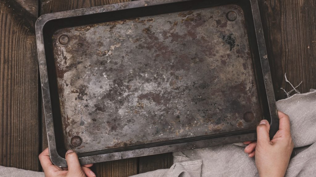 Two female hands hold a rectangular empty baking sheet on a brown wooden table, top view