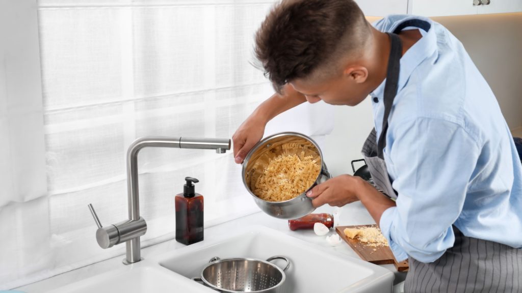 Man draining water from saucepan with pasta in messy kitchen