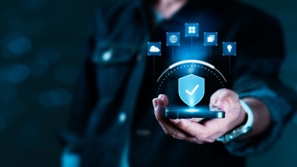 A person is holding a cell phone with a blue shield on it. The shield is surrounded by a blue circle with a checkmark inside. Concept of digital security, internet protection and cyber crime