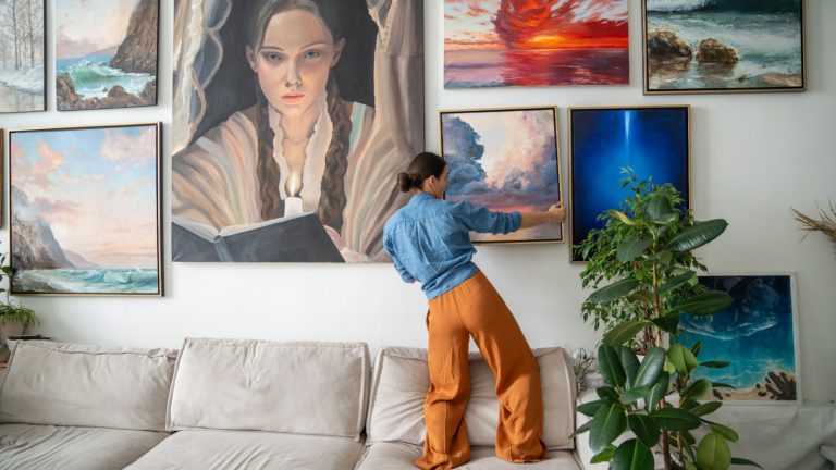 Interested woman painter, hangs on wall trendy artwork, created handmade in art workshop. Inspired pensive female artist designer holds new finished painting ponder compose for decoration art studio.