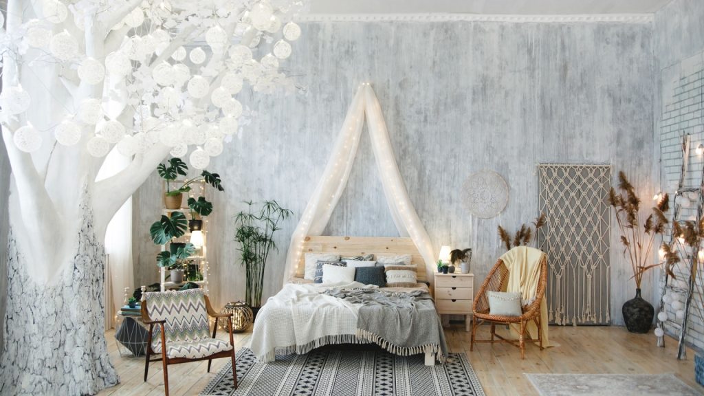 Elegant boho bedroom setup with white tree with cozy lights, soft bedding, woven chair, houseplants, light grey walls, and boho-chic accents. Room includes decorative rugs and artistic decor pieces