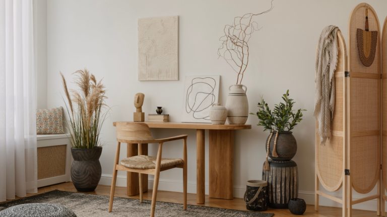 Modern home office with a minimalist design, featuring a wooden desk with decorative items, a chair, and potted plants. A doorway reveals a cozy adjoining room. Natural light and atmosphere.