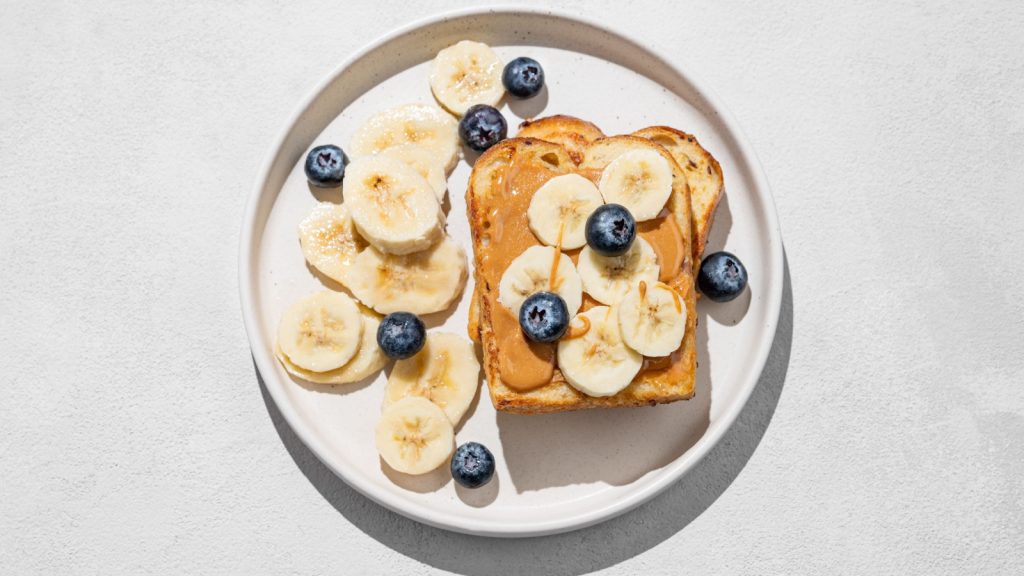 Peanut butter toast on a plate with banana and blueberry on a light background with  shadow. Concept of sweet dietary and healthy snacks for american breakfast. Top view and copy space.