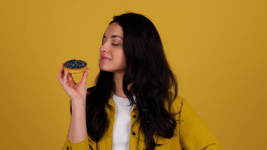 Young pretty lady biting a blueberry tartlet, posing in studio, isolated on yellow background. Food, lifestyle concept. Real time