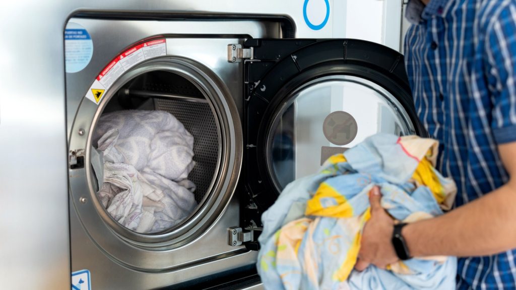Hotel worker loading a large capacity washing machine with bed sheets and towels, managing laundry in a professional setting