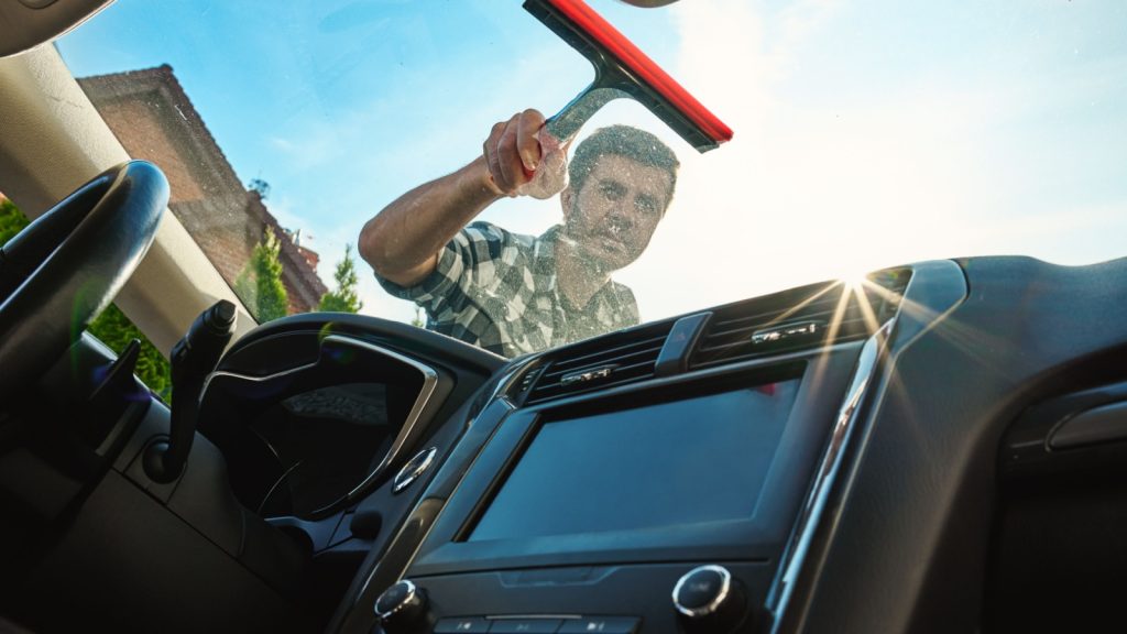 Male washing windshield in sunlight, view from from car inside. Man cleaning car windshield from outside using squeegee. Concept of car care, cleaning services and automotive hygiene