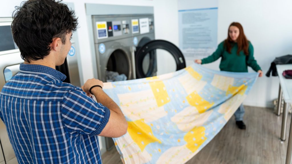 Young couple folding a clean bed sheet together after washing it in a public laundromat, highlighting sustainable cleaning practices and teamwork in housework