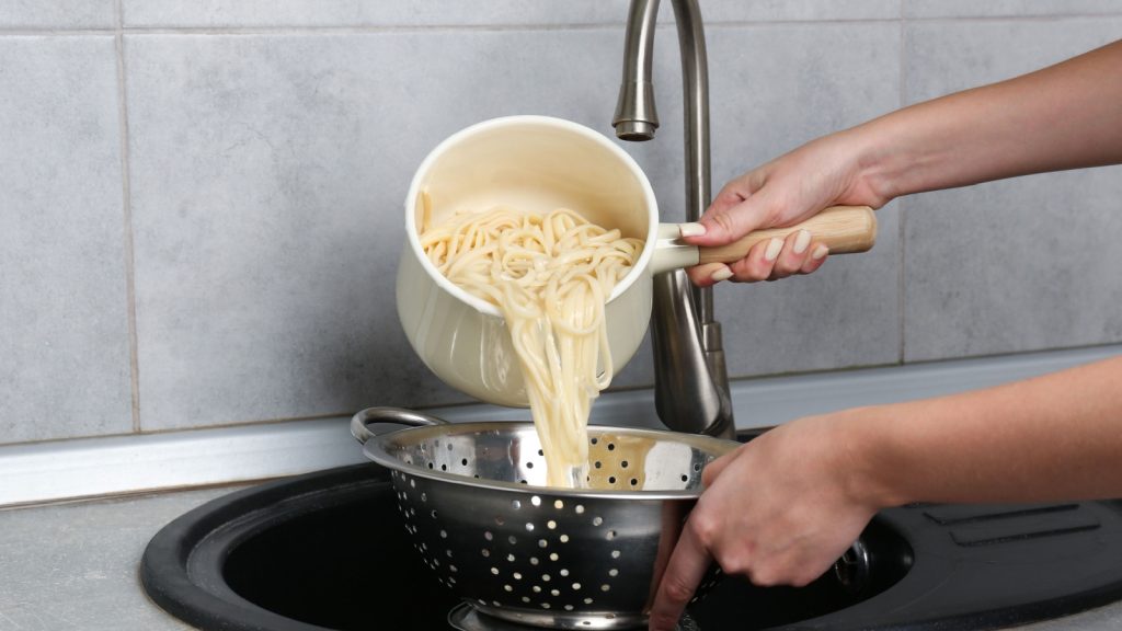 Woman draining boiled tagliatelle pasta into colander at sink in kitchen, closeup