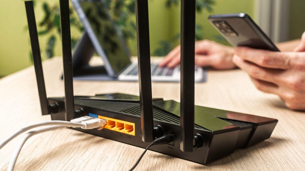 Wireless internet router on light office desk, user in background works on laptop and phone, configuring stable home network connection