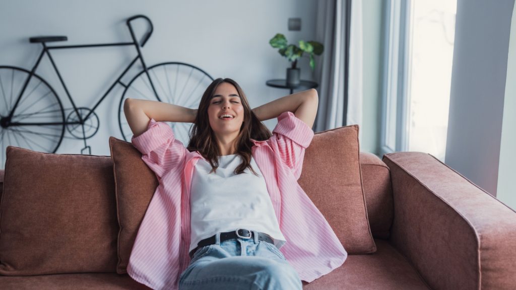 A relaxed young woman lounges on a comfortable sofa at home with her hands behind her head, feeling calm and happy, dreaming, enjoying wellbeing, and breathing fresh air in a cozy modern living room