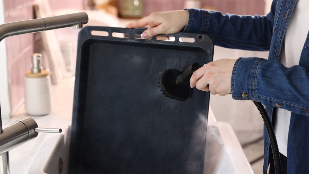 Woman cleaning baking sheet with steam cleaner in sink in kitchen, closeup