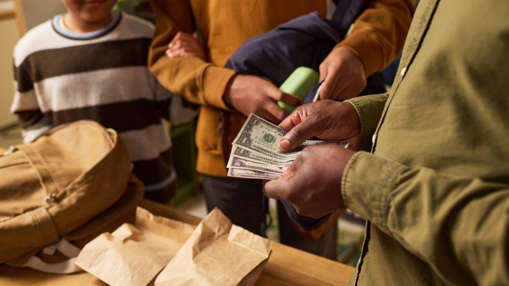 Black parent holding cash while children preparing school bags and lunch, representing family budgeting and education costs. Useful for back to school spending, household finance campaigns