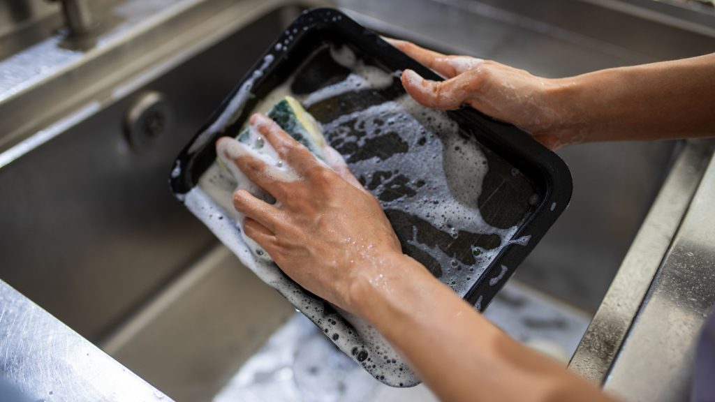 Close-up of hands washing a baking tray in a stainless steel sink, covered in soap suds, highlighting household chores and cleanliness.