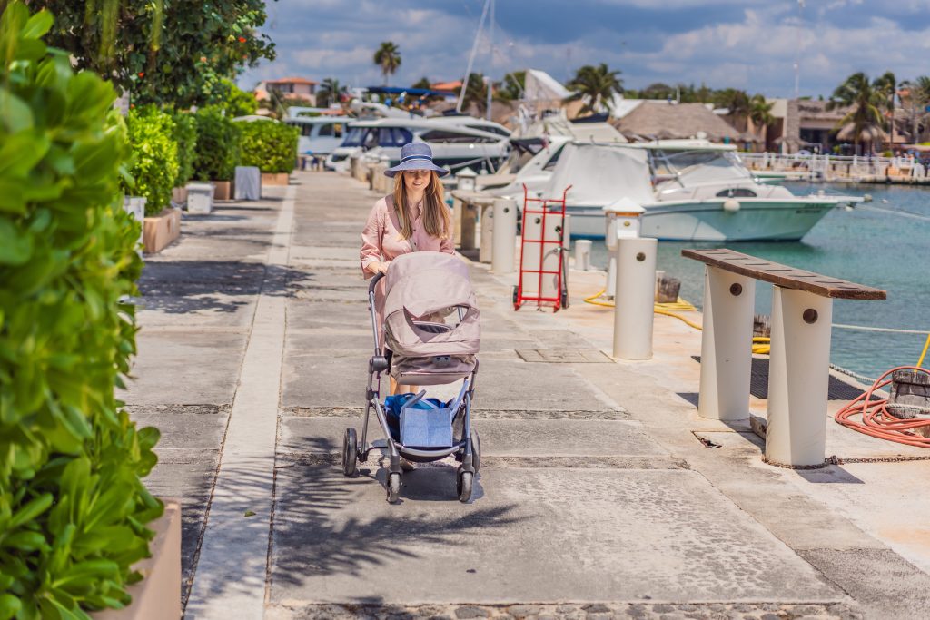 A young mother walks along the scenic waterfront, pushing a baby stroller with her child inside. A peaceful and heartwarming moment of motherhood, relaxation, and outdoor bonding by the sea.