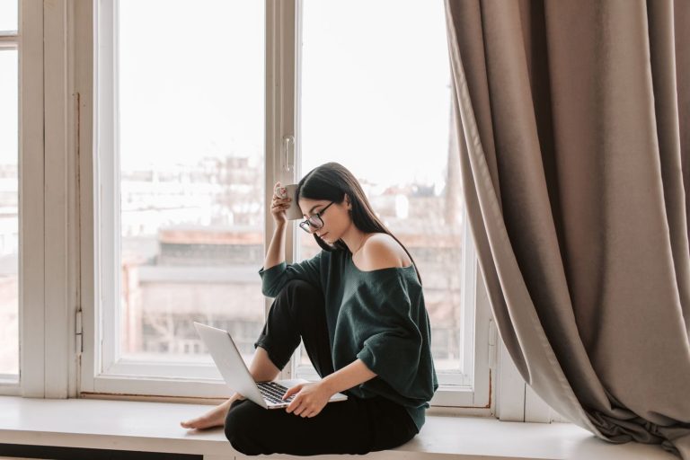 Side view full body of focused young lady in eyeglasses and dark sweater with open shoulder working on netbook while sitting barefoot on windowsill and enjoying cup of hot beverage