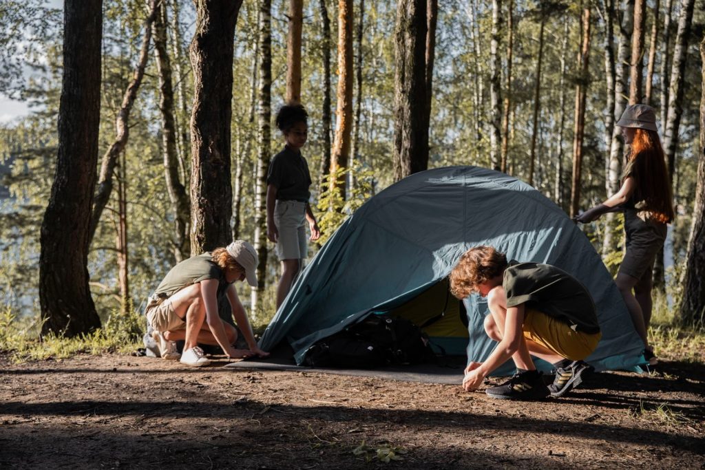 Group of teenagers setting up a tent during a camping trip in a sunlit forest.