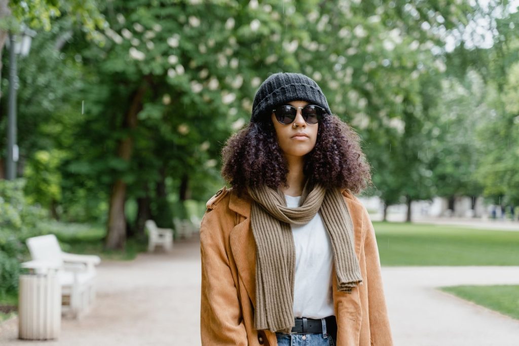 Young woman in a brown coat and sunglasses walking through a green park.