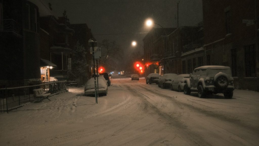 Snow-covered street with parked cars and red lights at night.