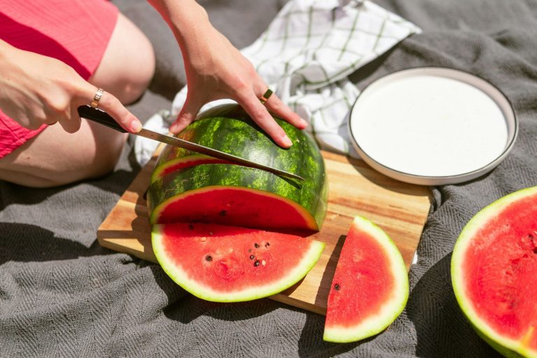 A person cutting a fresh watermelon on a picnic blanket.